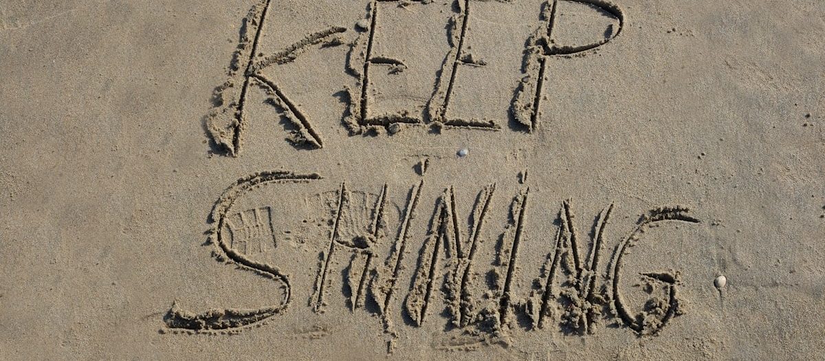 The words "KEEP SHINING" are carefully etched into the golden sand of a peaceful beach, their letters slightly uneven, as if written by hand with a gentle touch. The sunlight enhances the texture of the sand, casting soft shadows that give the message depth and warmth. Nearby, the rhythmic waves approach, their shimmering reflections reinforcing the idea of perseverance and radiance. The scene exudes positivity and motivation, as if nature itself is encouraging the viewer to keep shining, no matter the challenges ahead.