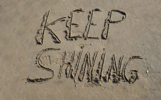 The words "KEEP SHINING" are carefully etched into the golden sand of a peaceful beach, their letters slightly uneven, as if written by hand with a gentle touch. The sunlight enhances the texture of the sand, casting soft shadows that give the message depth and warmth. Nearby, the rhythmic waves approach, their shimmering reflections reinforcing the idea of perseverance and radiance. The scene exudes positivity and motivation, as if nature itself is encouraging the viewer to keep shining, no matter the challenges ahead.