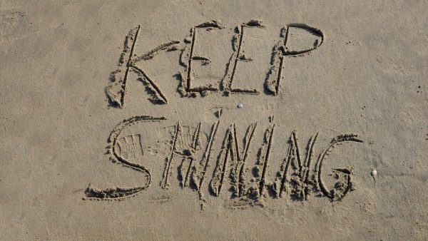 The words "KEEP SHINING" are carefully etched into the golden sand of a peaceful beach, their letters slightly uneven, as if written by hand with a gentle touch. The sunlight enhances the texture of the sand, casting soft shadows that give the message depth and warmth. Nearby, the rhythmic waves approach, their shimmering reflections reinforcing the idea of perseverance and radiance. The scene exudes positivity and motivation, as if nature itself is encouraging the viewer to keep shining, no matter the challenges ahead.