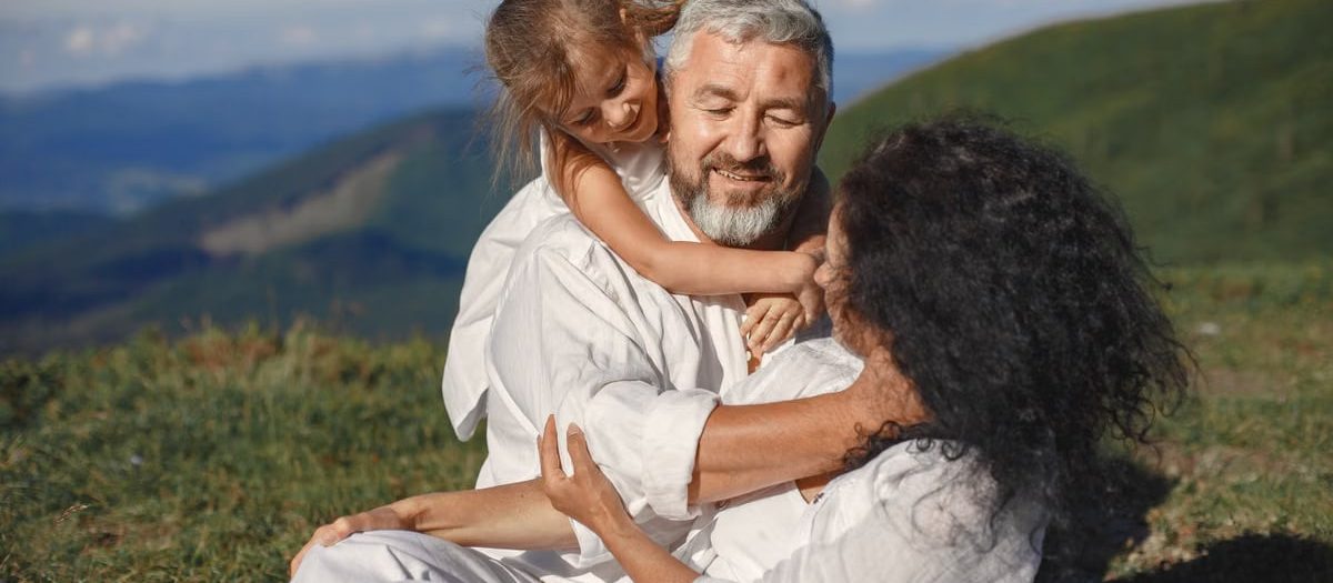 A joyful family moment outdoors, showing grandparents hugging their granddaughter under a sunny summer sky.
