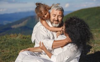 A joyful family moment outdoors, showing grandparents hugging their granddaughter under a sunny summer sky.