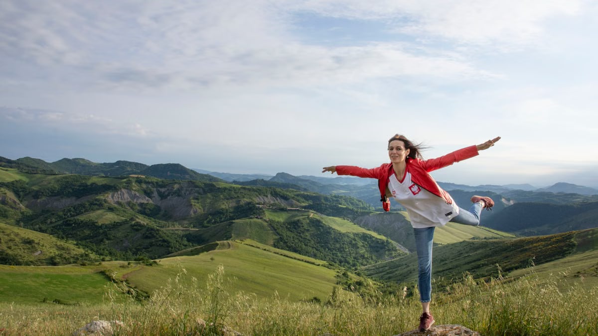 A woman joyfully balances on a rock amidst rolling green hills under a clear blue sky.